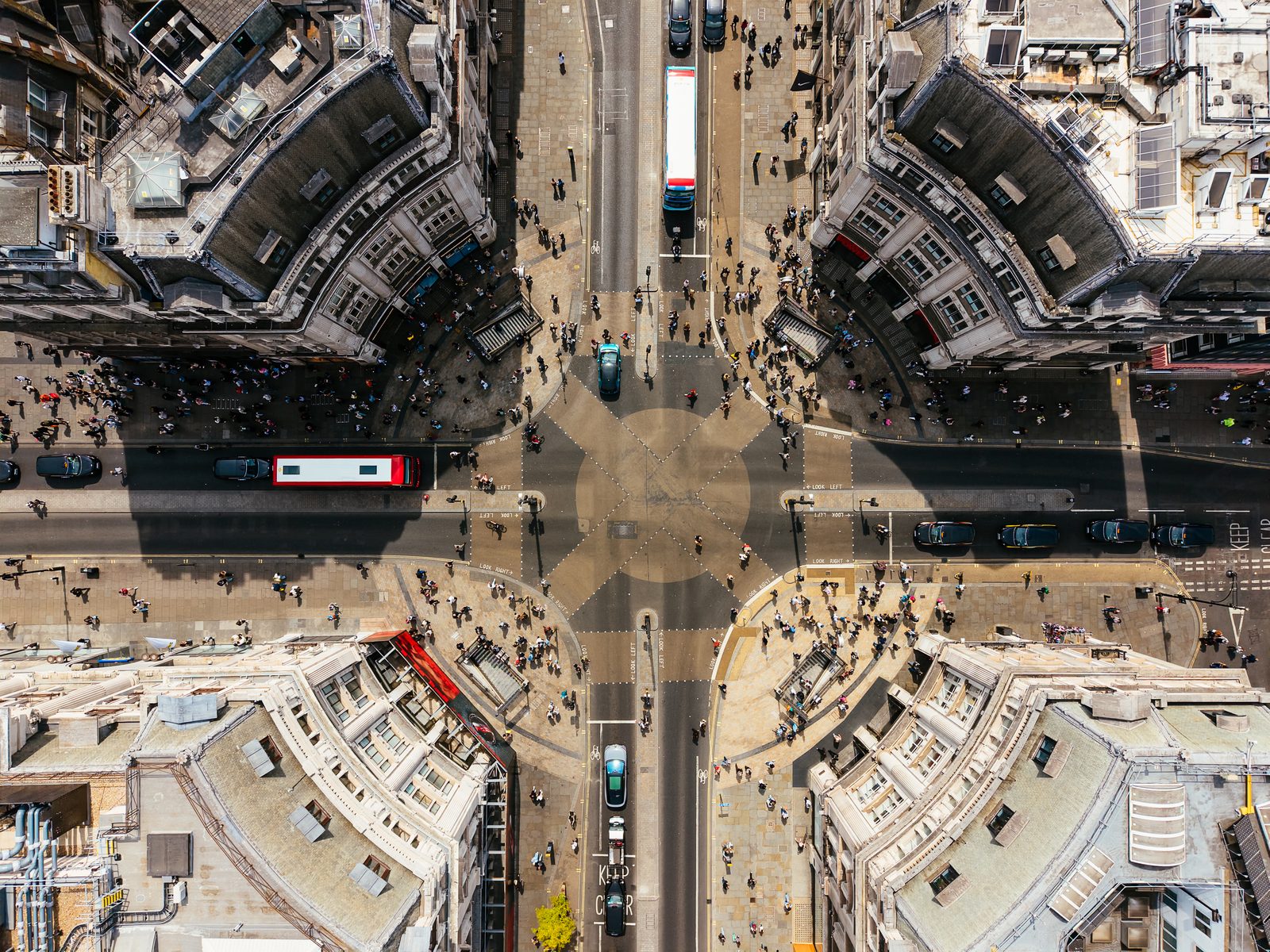 A busy London street crossing seen from directly overhead, buses, taxis and crowds of people moving across it.