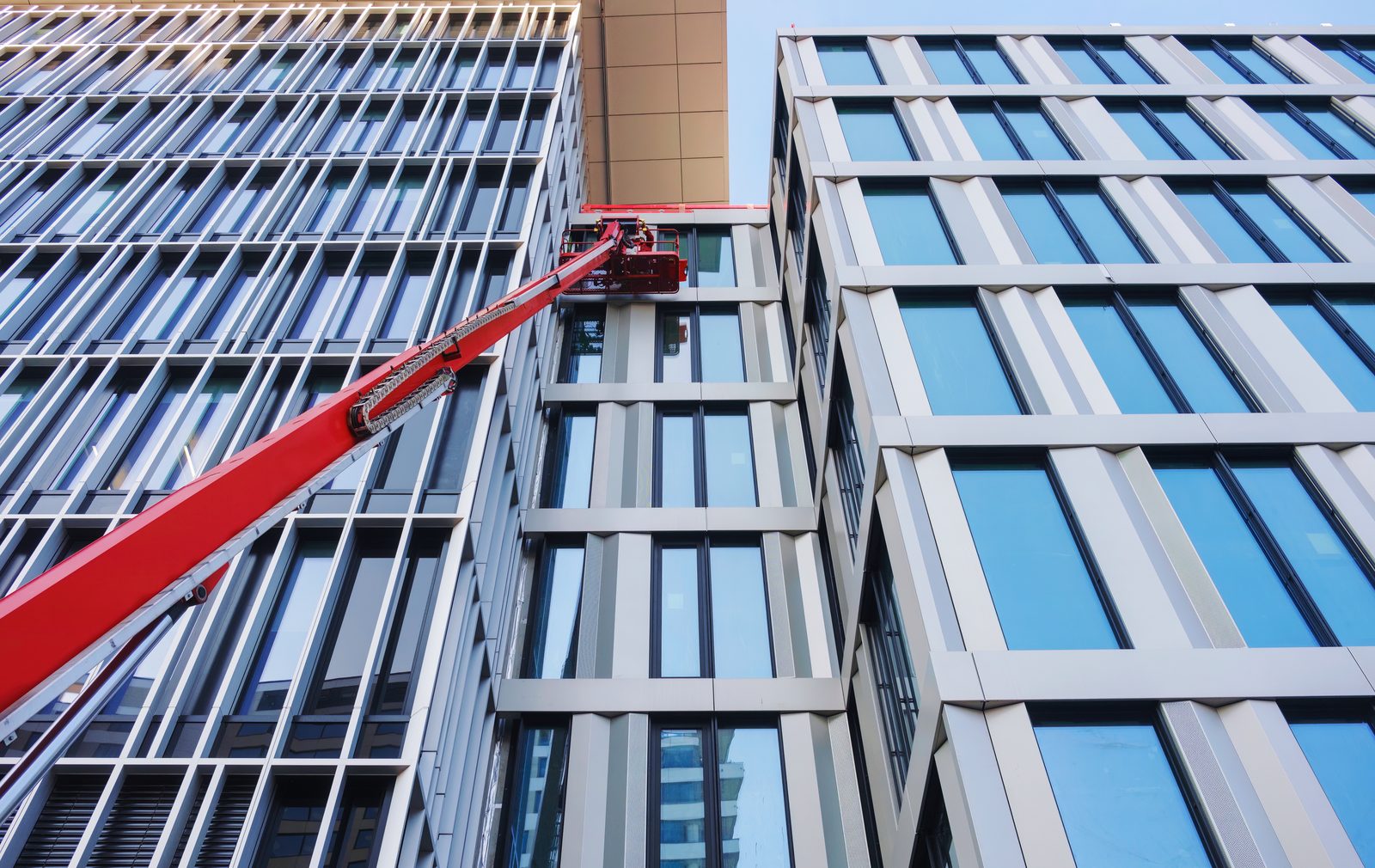 The red boom of a cherry picker reaching up to a working platform against the glazed facade of a building.