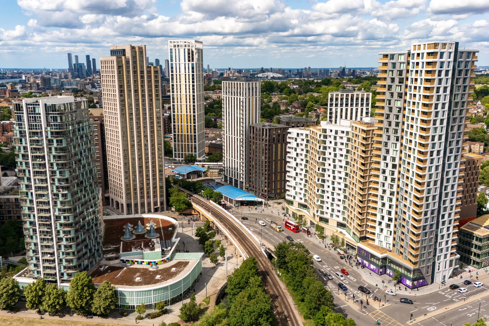An aerial view over a London district, residential towers around a station and a high street, with the wider city beyond.